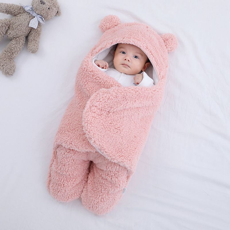 Baby wrapped in a pink fluffy swaddle with bear ears, next to a teddy bear on a white background