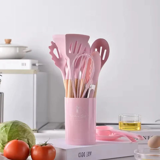 Pink kitchen utensil set in a holder on a kitchen counter with vegetables and a blender in the background.